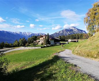 Eine malerische Almlandschaft mit einem Bauernhaus und hohen Bergen im Hintergrund. Der Weg führt durch die grüne Wiese unter einem klaren Himmel.