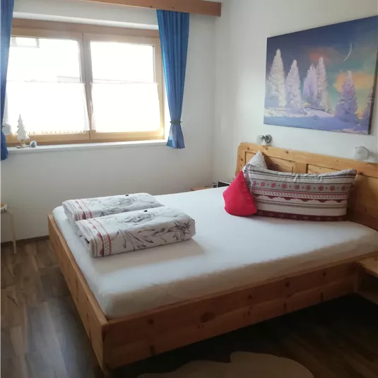 Ein gemütliches Schlafzimmer mit einem Holzbett und bunten Kissen. Große Fenster lassen viel Licht herein und sorgen für eine helle Atmosphäre.