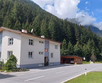A modern building in a green environment with mountains in the background. The road in front of the house is well-traveled and there is plenty of nature all around.