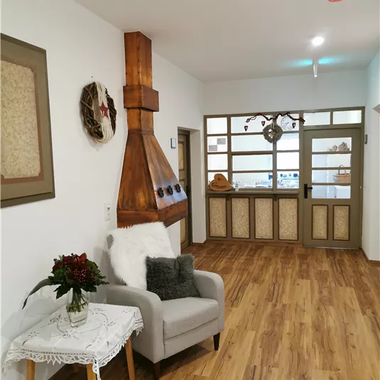 A bright room with a cozy armchair and a decorative table. In the background, a wood stove and a glass door can be seen.