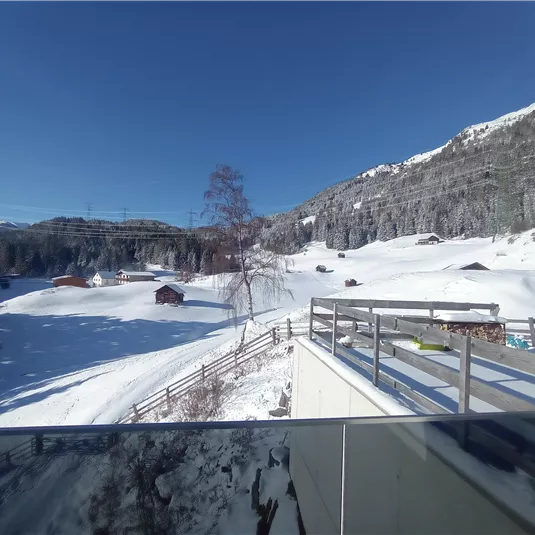 A snowy landscape with small cottages and a clear blue sky. In the background, mountains and trees can be seen.