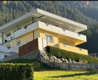 Ein modernes Haus auf einem Hügel, umgeben von Grünflächen. Im Hintergrund sind Wald und Berge sichtbar.