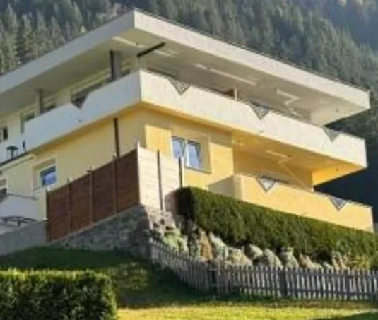 A modern house on a hill, surrounded by green spaces. In the background, forest and mountains are visible.