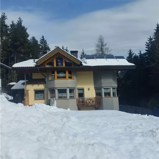 Ein modernes Haus mit einem großen Schneehaufen davor. Im Hintergrund sind Bäume und ein klarer Himmel zu sehen.