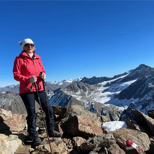 A woman stands on a peak in the mountains wearing a red jacket. In the background, there are snow-covered mountains and a clear sky.
