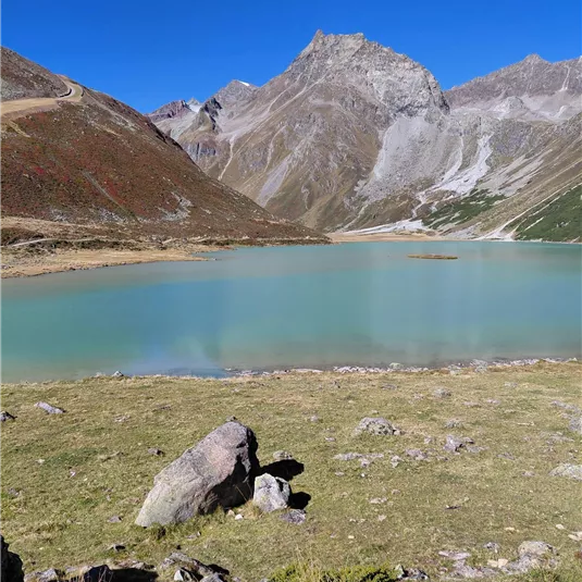 A quiet, blue mountain lake surrounded by impressive mountains and green grass. The sky is clear and sunny.