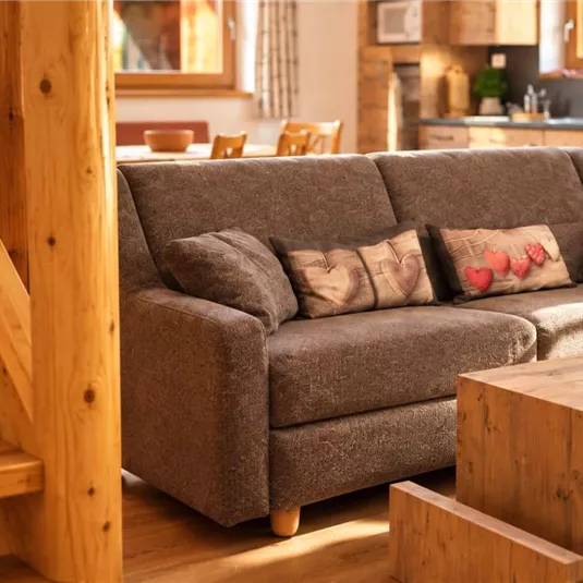 Ein gemütliches Wohnzimmer mit einem grauen Sofa und Holzdekor. Im Hintergrund sind eine Treppe und eine moderne Küche zu sehen.