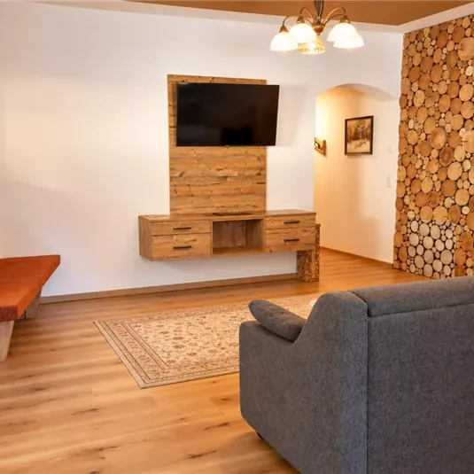 Ein gemütliches Wohnzimmer mit einem grauen Sofa und einem Fernseher an der Wand. Die Holzvertäfelung und der Holzkamin verleihen dem Raum eine warme Atmosphäre.