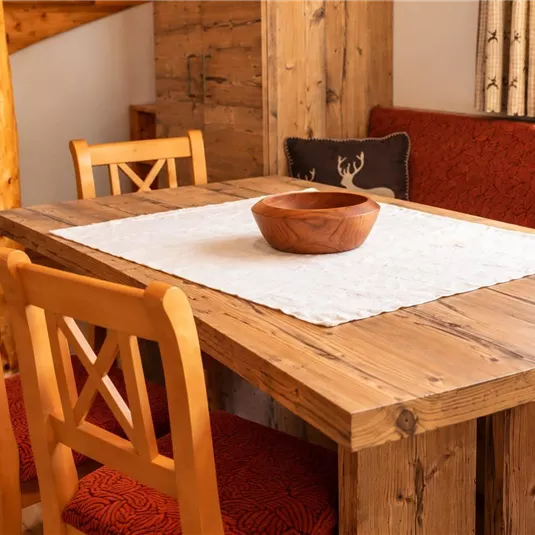Ein holzfarbenes Esstisch mit vier Stühlen steht in einem gemütlichen Raum. In der Mitte des Tischs befindet sich eine Holzschüssel auf einem weißen Tischläufer.