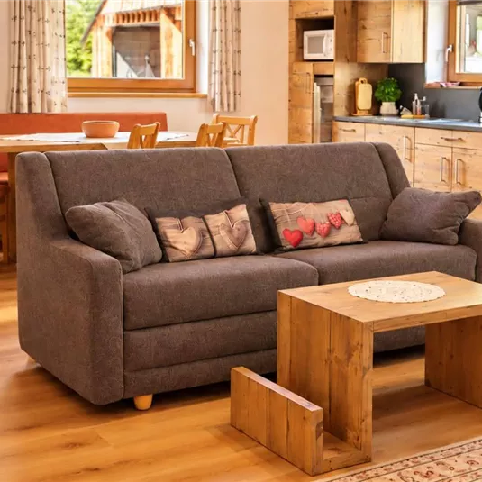 Ein modernes Wohnzimmer mit einem grauen Sofa und einem hölzernen Tisch. Die Küche im Hintergrund ist gut ausgestattet, und der Raum hat viel natürliches Licht.