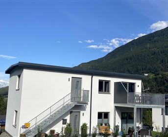 A modern two-story house with an outdoor staircase. In the background, green mountains and a blue sky can be seen.