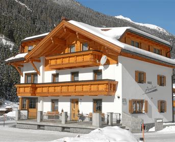 A cozy chalet in the snow with wooden cladding and several balconies. The surrounding mountains are covered with a thick layer of snow.