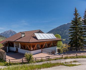Ein modernes Haus mit einer Holzveranda und Solarpanelen auf dem Dach. Im Hintergrund sind Berge und grüne Wiesen zu sehen.