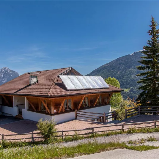 A modern house with a wooden porch and solar panels on the roof. In the background, mountains and green meadows can be seen.