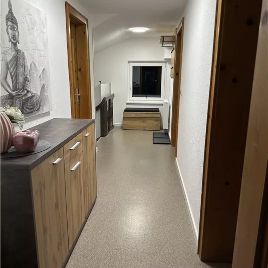 A bright hallway with a wooden table and a Buddha representation on the wall. At the end of the hallway, there is a window and a seating area.