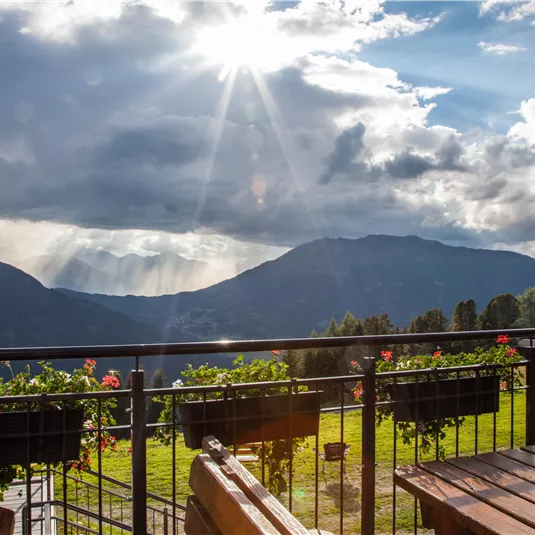 Ein schöner Ausblick auf die Berge mit strahlendem Sonnenlicht und dramatischen Wolken. Blühende Pflanzen und Holzstühle im Vordergrund verleihen der Szene eine einladende Atmosphäre.