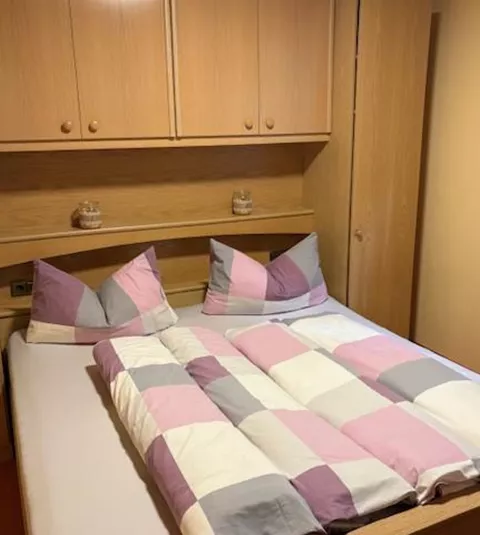 Ein gemütliches Schlafzimmer mit einem Doppelbett und bunten Kissen. Der Raum ist einfach und hell mit Holzregalen.