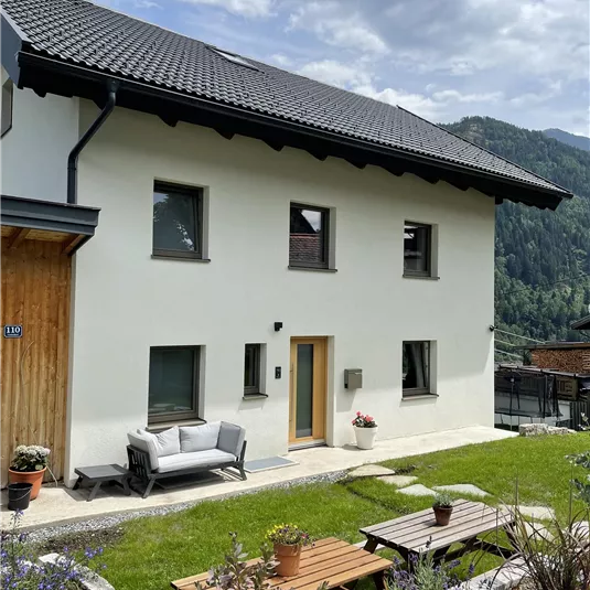 A modern house with a well-maintained garden. In the background, green mountains and a clear sky are visible.