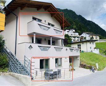 Ein modernes Gebäude mit einem Balkon und einer gemütlichen Terrasse. Die Umgebung ist von grünen Bergen und anderen Häusern umgeben.