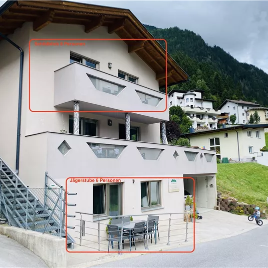 A modern building with a balcony and a cozy terrace. The surroundings are surrounded by green mountains and other houses.