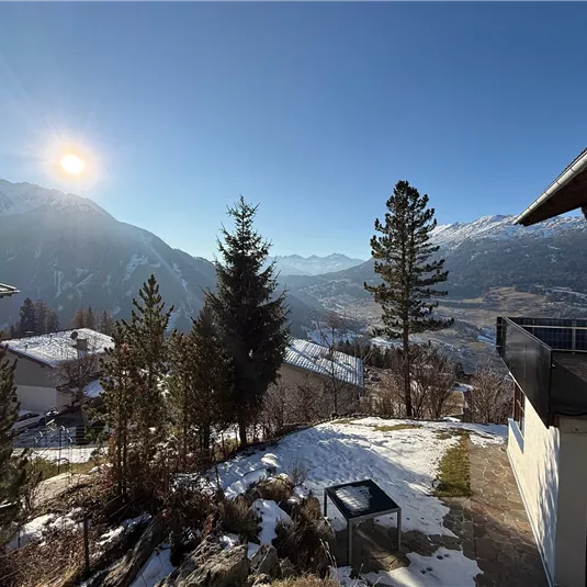 A beautiful mountain landscape with snow-covered hills and radiant sunshine. In the foreground, there are some trees and a modern building.