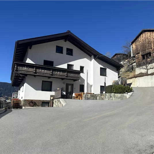 Ein modernes Haus mit einem großzügigen Parkplatz, umgeben von Bergen. Im Hintergrund befinden sich traditionelle Holzgebäude.