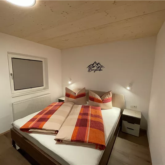 A cozy bedroom with a large bed and colorful pillows. The walls are bright, and there is a window with a simple curtain.