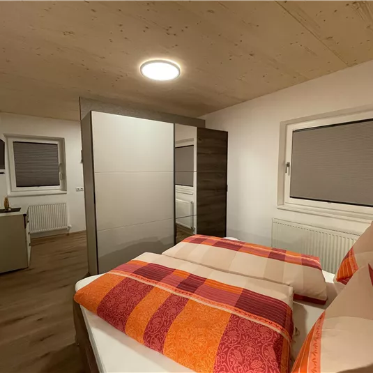 A modern bedroom with two beds and colorful cushions. The room has a bright atmosphere with wooden and gray accents.