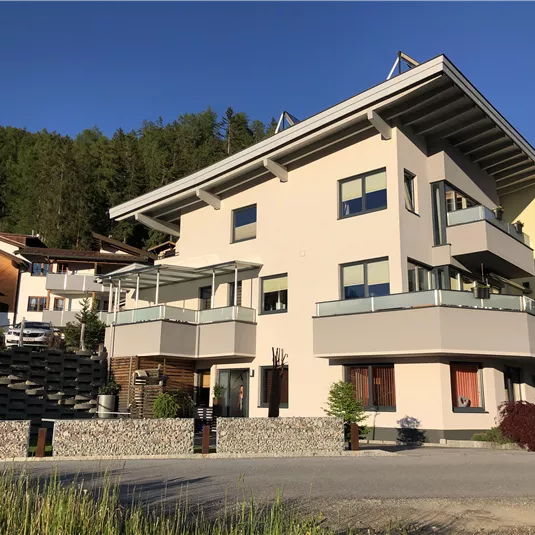 Ein modernes Gebäude mit mehreren Balconen in einer ruhigen Umgebung. Im Hintergrund sind weitere Wohnhäuser und Bäume zu sehen.