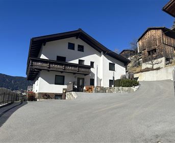 A modern house with a spacious parking lot, surrounded by mountains. Traditional wooden buildings are in the background.