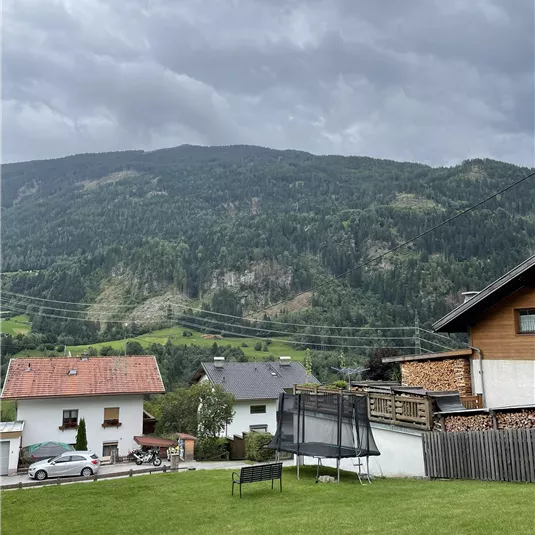 A picturesque landscape with hills and trees in the background. In the foreground, houses and a garden can be seen.