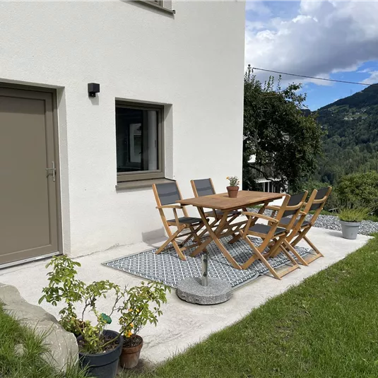 A cozy outdoor area with a wooden table and chairs. In the background, green hills and a blue sky can be seen.