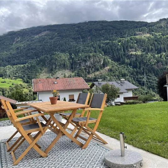 A cozy terrace with a wooden table and chairs. In the background are green hills and houses in a picturesque landscape.