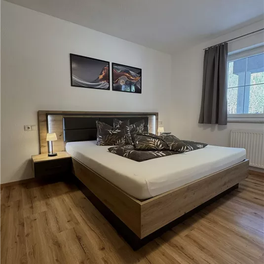 A modern bedroom with a large bed and decorative pillows. On the wall hang two pictures and a window provides daylight.