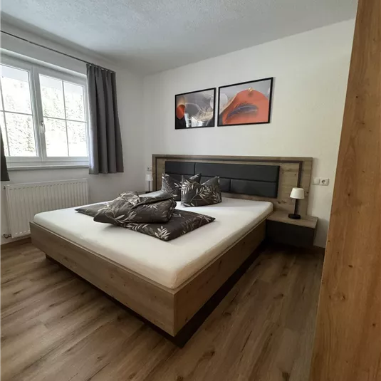 A modern bedroom with a large double bed and stylish pillows. The walls are light, and there are artworks and windows that let in plenty of daylight.