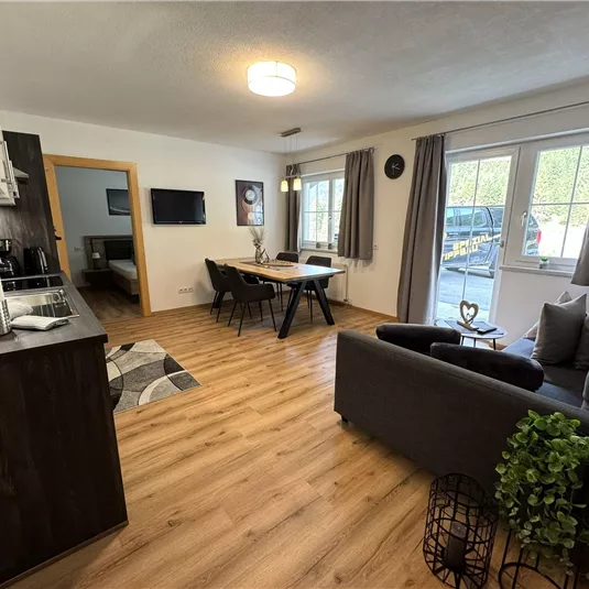 A modern furnished apartment with an open kitchen, dining table, and cozy living room. Large windows provide plenty of daylight and a view outside.