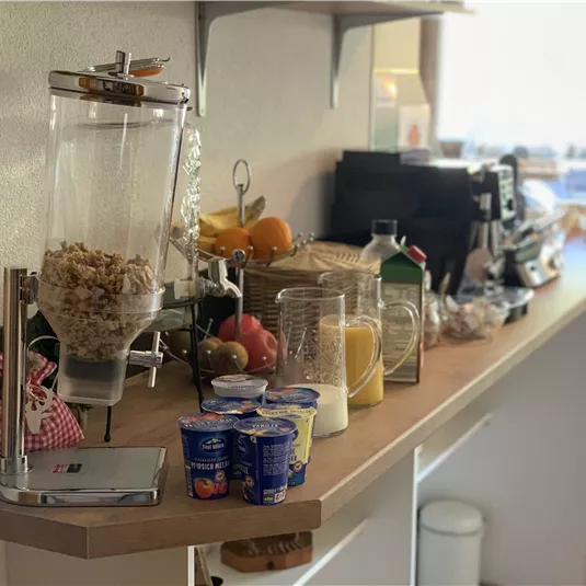 A modern kitchenette with a selection of breakfast ingredients. On display are cereals, fruit, yogurt, and drinks.