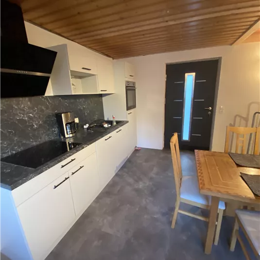 A modern kitchen with white cabinets and a dark countertop. There is a dining table with chairs and an entrance door in the background.