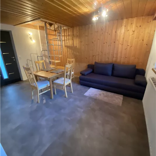 A cozy living room with a gray sofa and a wooden table. A wooden ceiling and a spiral staircase give the space a rustic charm.