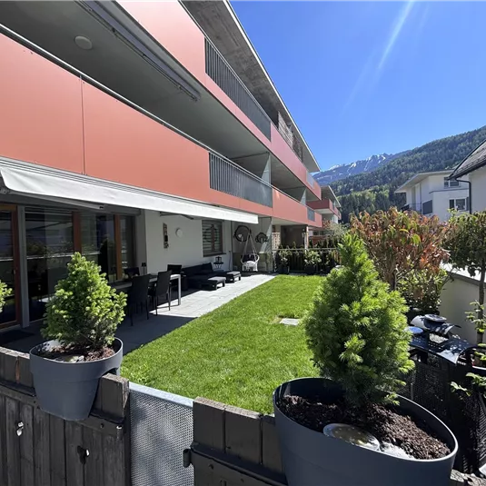 A modern residential building with a green lawn and patio furniture. The surroundings are sunny, with mountains in the background.