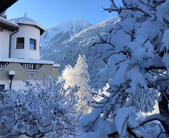 A snow-covered landscape with a cozy house and snow-covered trees. In the background, majestic mountains and a blue sky can be seen.