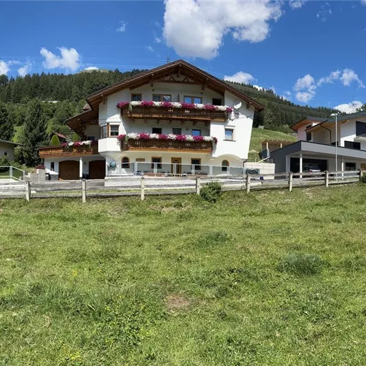 A farmhouse in nature with green meadows and trees in the background. The sky is blue and has some clouds.