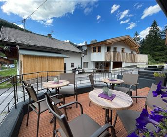 A cozy terrace with round tables and chairs. In the background, alpine houses and a clear sky are visible.