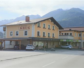 Ein Gasthaus mit der Aufschrift "KRUG" und einer gelben Fassade. Im Hintergrund sind Berge zu sehen und es stehen einige Autos vor dem Gebäude.