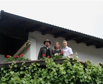 Three people are standing on a balcony, surrounded by blossoming greenery. They are wearing festive clothing and appear cheerful.