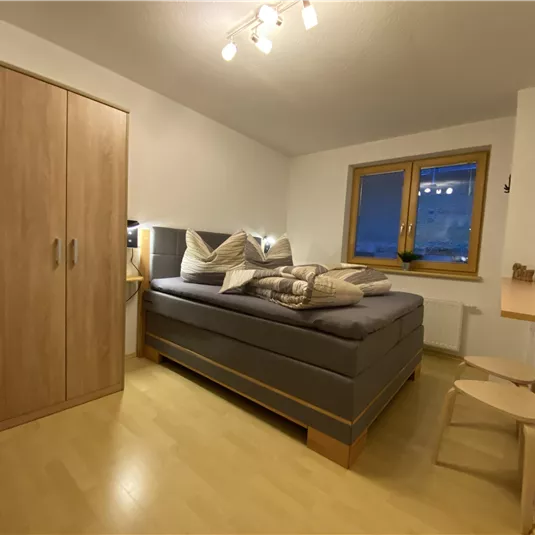 A cozy bedroom with a large bed and a wooden wardrobe. The light is soft, and there is a window overlooking the outside.