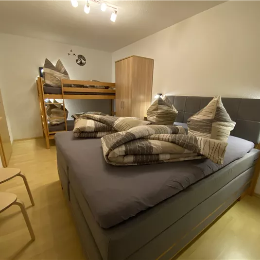 A cozy bedroom with a large double bed and a bunk bed. The decor is modern and minimalist, featuring light wood and neutral colors.