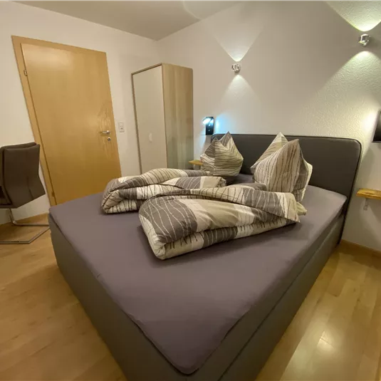 A modern bedroom with a large bed and a desk. The walls are light and there are warm wood details.