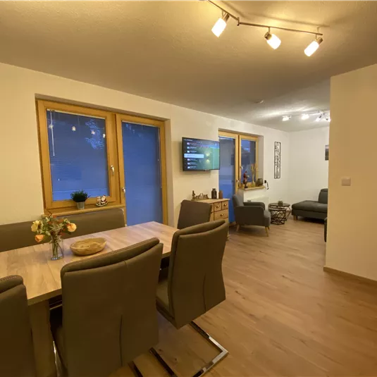 A modern living room with a dining table and chairs. Large windows let in plenty of light and the room is cozily furnished.