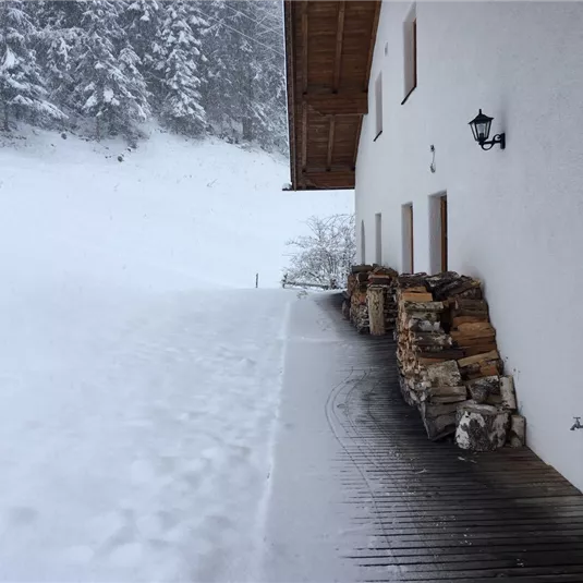 A snow-covered landscape next to a building. Stacked logs are on the terrace.